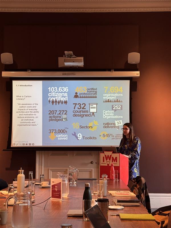 A woman stands at the end of a table, behind a red stand with 'IWM' on it. She is in front of a presentation displaying some key numbers around carbon literacy training